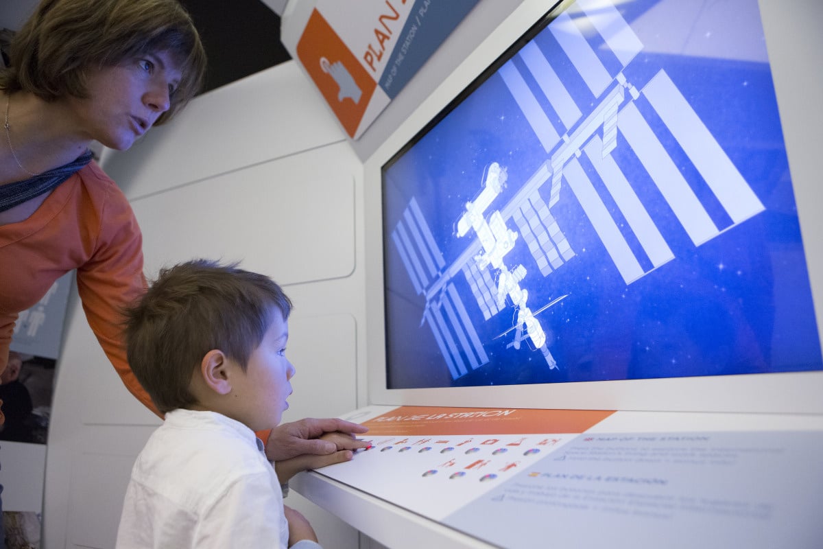 Une mère et son enfant appuient sur un bouton du plan intéractif de la station spatiale dans Astronautes l'expo